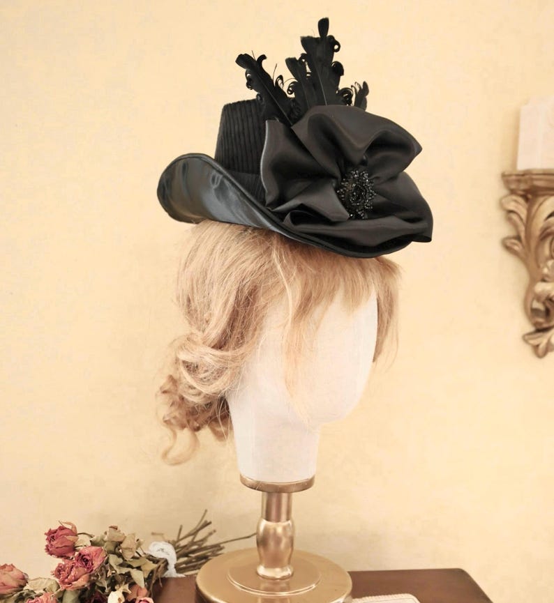 May include: A black hat with a wide brim and a large black bow on top. The hat is decorated with black feathers and a small black jewel.