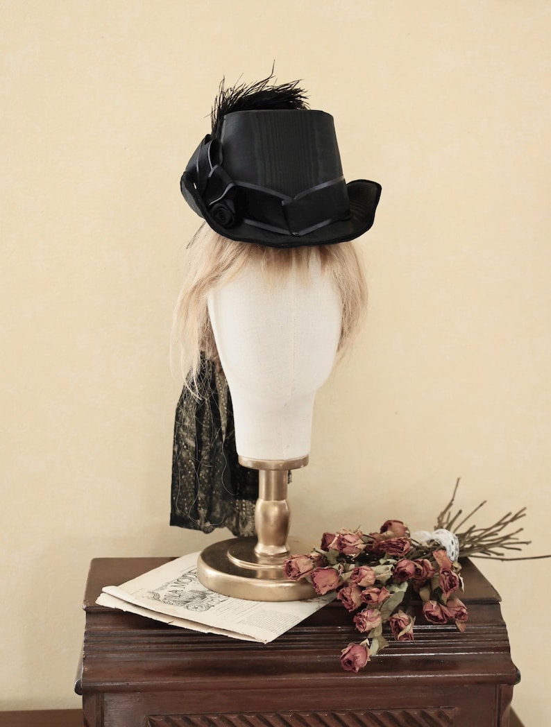 Op de afbeelding: Een zwarte hoge hoed met een zwarte veer en een zwart lint. De hoed zit op een wit mannequin hoofd.