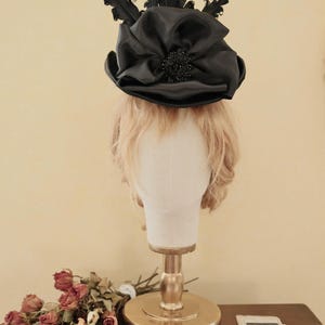 May include: A black satin top hat with a large bow and black feathers. The hat is on a mannequin head with blonde hair.