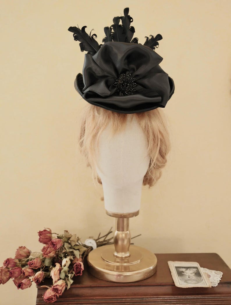 May include: A black satin top hat with a large bow and black feathers. The hat is on a mannequin head with blonde hair.