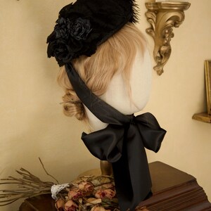 May include: A black hat with a floral design and feather trim, secured with a black satin ribbon. The hat is displayed on a mannequin head with blonde hair, and is placed on a wooden surface.