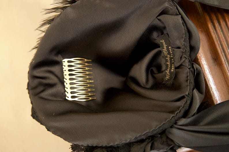 May include: A black satin headpiece with a gold comb and feather trim. The label reads "Melancholy Chapel, Made in France". The headpiece is likely vintage and suitable for special occasions.