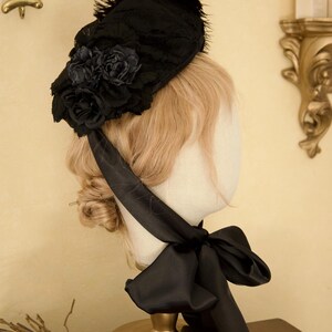 May include: A black lace bonnet with a cluster of dark roses and feather accents. A black satin ribbon is tied in a bow under the chin. The bonnet is displayed on a mannequin head with blonde hair.