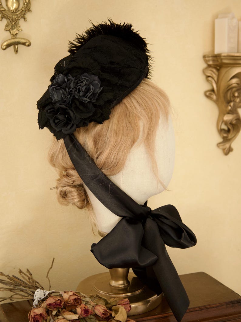 May include: A black lace bonnet with a cluster of dark roses and feather accents. A black satin ribbon is tied in a bow under the chin. The bonnet is displayed on a mannequin head with blonde hair.