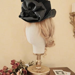 May include: A black top hat with a large black satin bow and black feathers. The hat is on a white mannequin head with blonde hair.