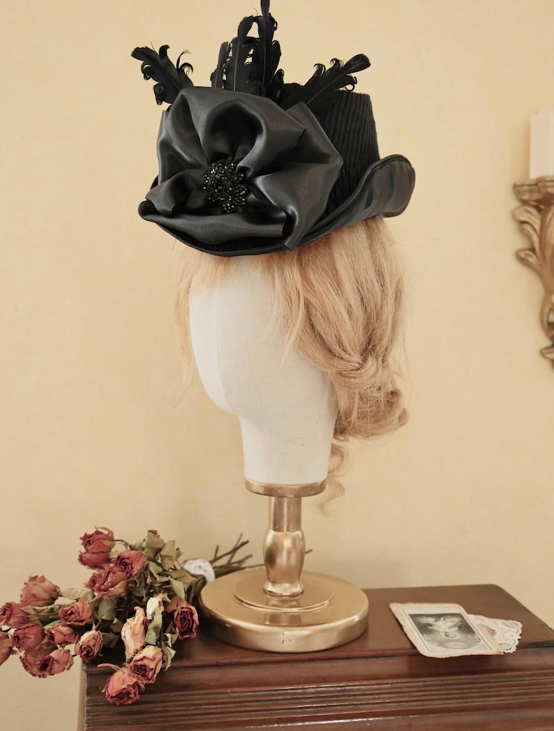 May include: A black top hat with a large black satin bow and black feathers. The hat is on a white mannequin head with blonde hair.