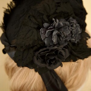May include: A black vintage hat with a floral design. The hat features a lace overlay and is adorned with dark gray fabric flowers. A black ribbon is attached to the hat. The hat is likely from the Victorian or Edwardian era.