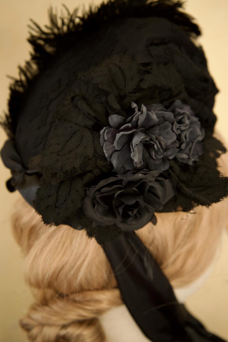 May include: A black vintage hat with a floral design. The hat features a lace overlay and is adorned with dark gray fabric flowers. A black ribbon is attached to the hat. The hat is likely from the Victorian or Edwardian era.