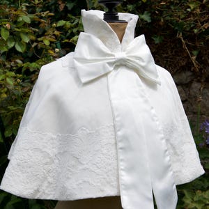 Peut inclure: Une cape de mariée blanche avec un grand nœud au cou. La cape présente une bordure en dentelle et une finition satinée. La cape est présentée sur un mannequin devant un fond de verdure.