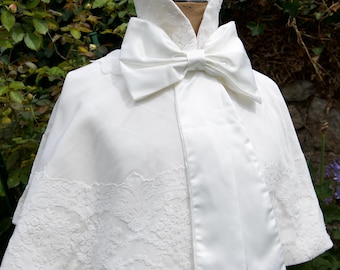 May include: A white bridal cape with a large bow at the neck. The cape features a lace trim along the bottom edge and a satin finish. The cape is displayed on a mannequin against a backdrop of green foliage.
