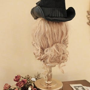 May include: A black velvet top hat with a wide brim and a black feather accent. The hat is sitting on a mannequin head with blonde curly hair.