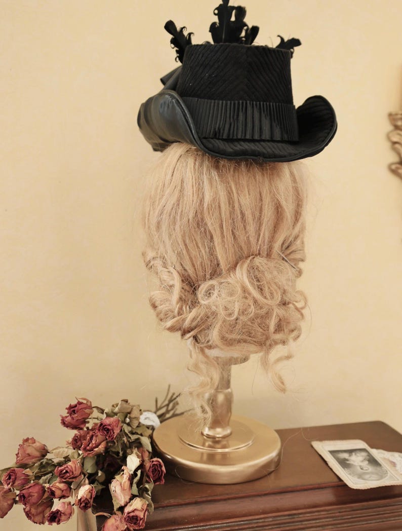 May include: A black velvet top hat with a wide brim and a black feather accent. The hat is sitting on a mannequin head with blonde curly hair.