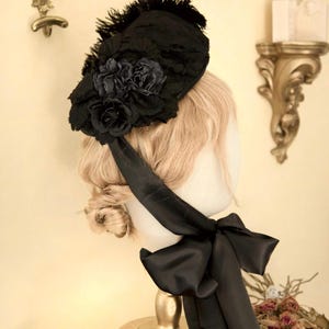 May include: A black hat with a floral design and feathers. The hat has a large black bow and is displayed on a mannequin head. The hat is made of lace and satin.