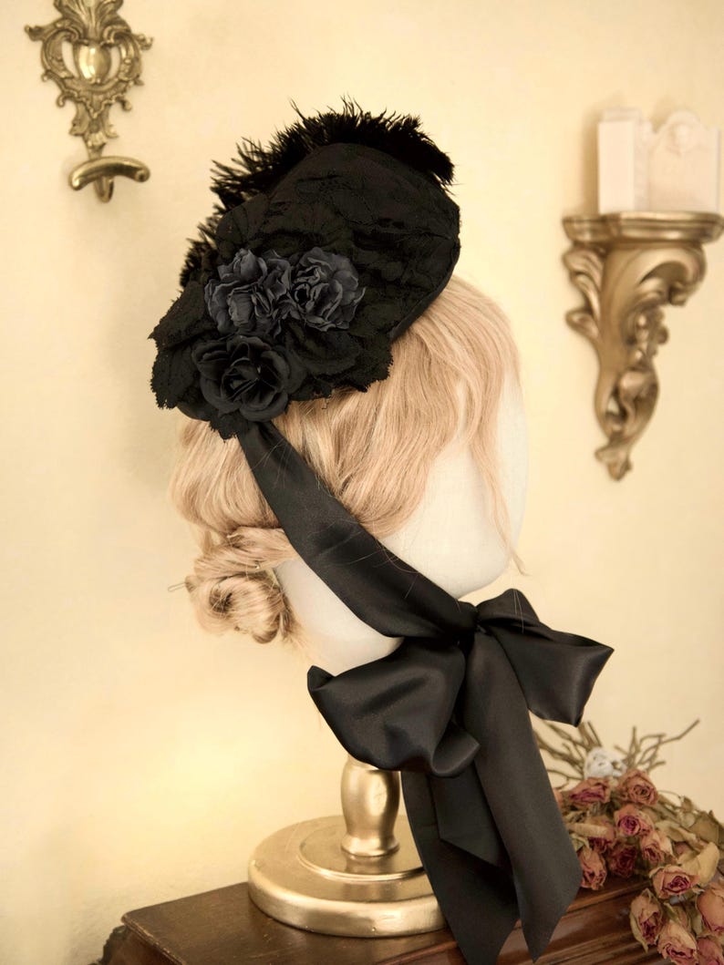 May include: A black hat with a floral design and feathers. The hat has a large black bow and is displayed on a mannequin head. The hat is made of lace and satin.