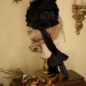 May include: A black vintage hat with floral and feather accents, displayed on a mannequin head. The hat features a large bow and ribbon detail. The background includes a wooden surface and decorative wall fixtures.