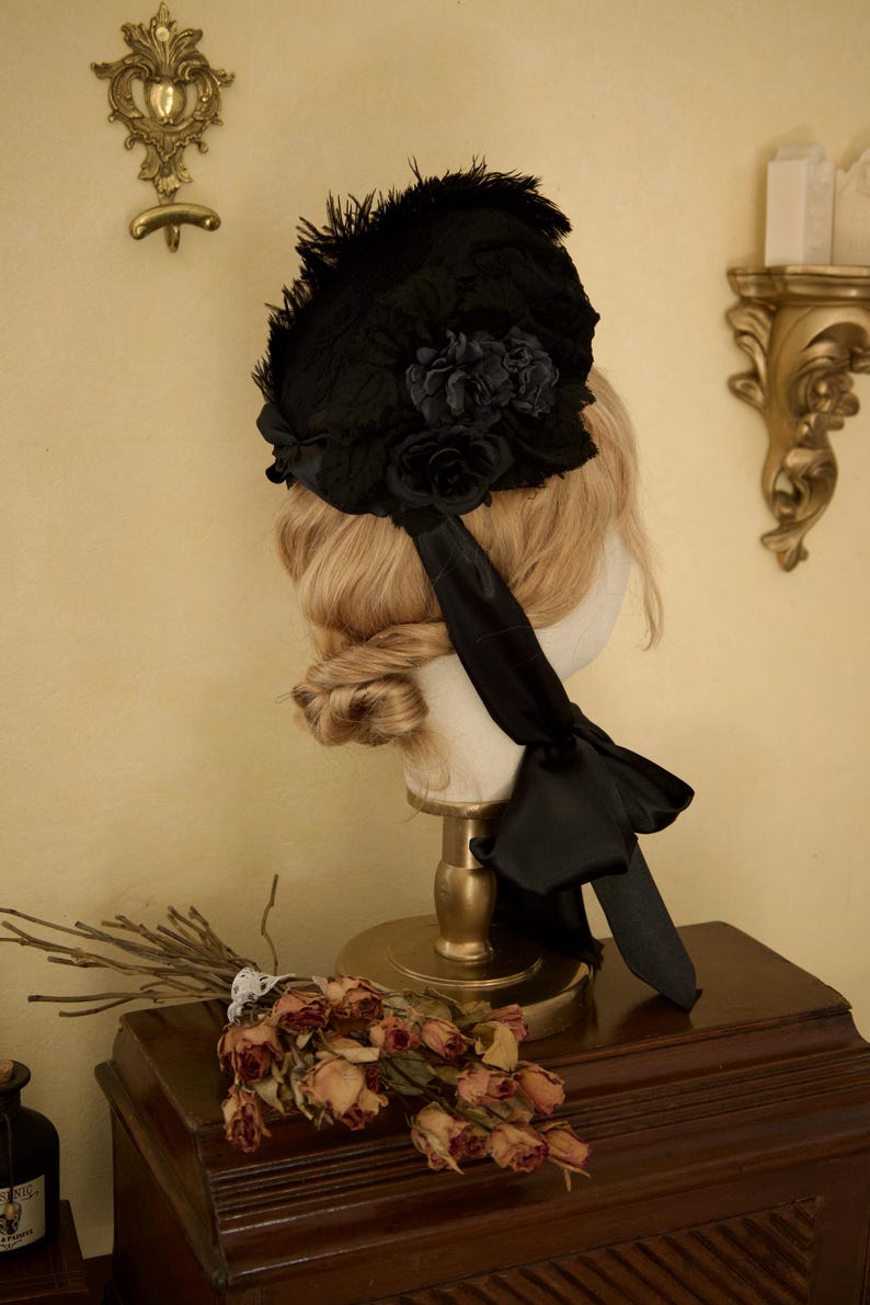 May include: A black vintage hat with floral and feather accents, displayed on a mannequin head. The hat features a large bow and ribbon detail. The background includes a wooden surface and decorative wall fixtures.