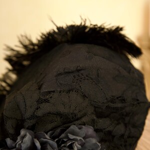 May include: Black lace hat with a dark feather trim and a cluster of dark fabric flowers. The hat's lace has a delicate floral pattern. The overall design is elegant and vintage-inspired, suitable for special occasions.