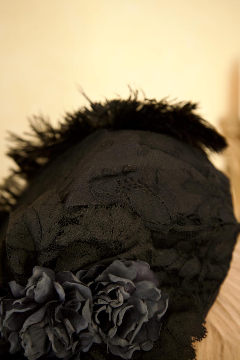 May include: Black lace hat with a dark feather trim and a cluster of dark fabric flowers. The hat's lace has a delicate floral pattern. The overall design is elegant and vintage-inspired, suitable for special occasions.