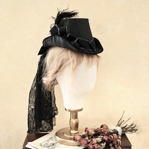 Op de afbeelding: Een zwarte hoge hoed met een zwarte kant sluier en een veren accent. De hoed is op een mannequin hoofd met blond haar.