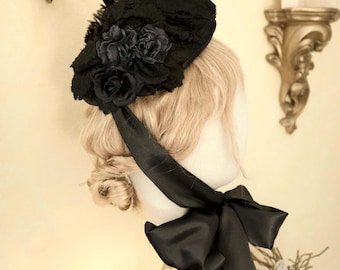 May include: A black hat with a floral design and feather accents. The hat features a large black bow and is displayed on a mannequin head. The hat is a vintage style.