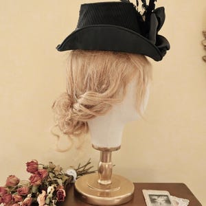 May include: A black hat with a wide brim and a decorative feather accent. The hat is on a mannequin head with blonde hair.