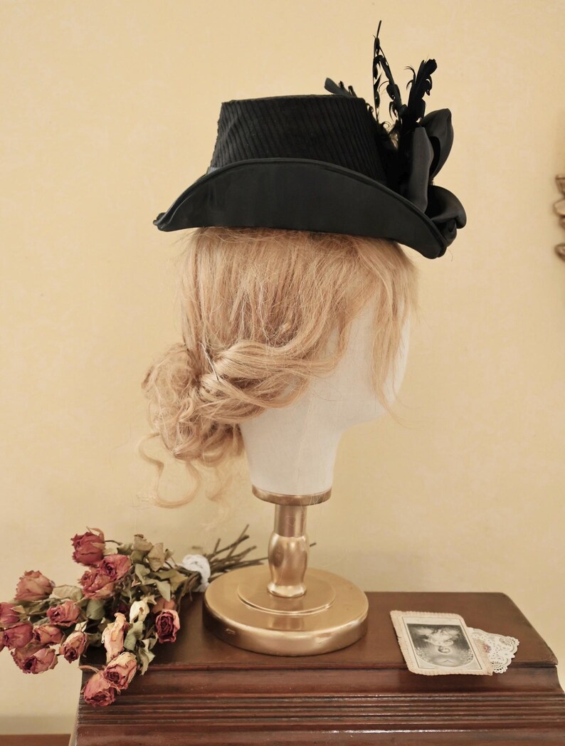 May include: A black hat with a wide brim and a decorative feather accent. The hat is on a mannequin head with blonde hair.