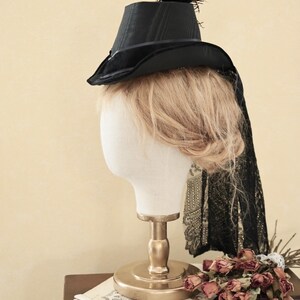 Op de afbeelding: Een zwarte fluwelen hoge hoed met een kleine zwarte veer bovenop. De hoed zit op een wit mannequin hoofd met blond haar. Het mannequin staat op een gouden standaard.
