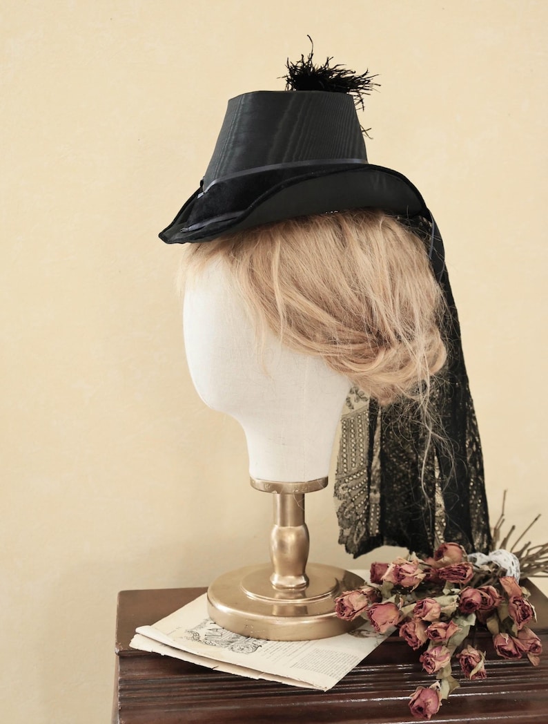 Op de afbeelding: Een zwarte fluwelen hoge hoed met een kleine zwarte veer bovenop. De hoed zit op een wit mannequin hoofd met blond haar. Het mannequin staat op een gouden standaard.