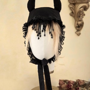 Könnte beinhalten: Ein schwarzer Teufelshut mit Spitzenbesatz und Perlenfransen. Der Hut hat zwei gebogene Hörner und eine schwarze Schleife. Das Kostümzubehör wird auf einem Schaufensterkopf mit blondem Haar präsentiert.