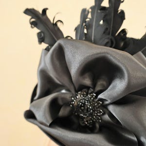 May include: A black satin hat with a large bow and black feathers. The bow has a black beaded embellishment in the center.