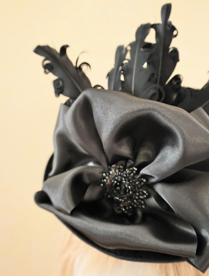 May include: A black satin hat with a large bow and black feathers. The bow has a black beaded embellishment in the center.