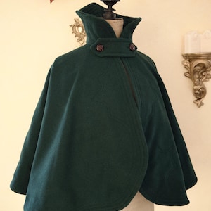 May include: A dark green cape with a high collar and two brown buttons. The cape is made of a thick, textured fabric and has a flowing design. The cape is displayed on a mannequin.