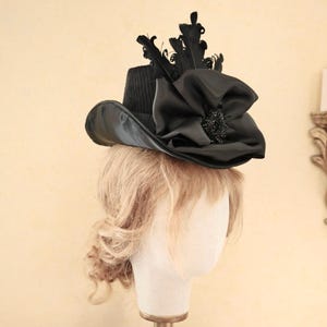 May include: A black hat with a wide brim and a large black bow on top. The hat is decorated with feathers and a black jewel.