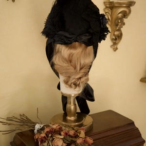 May include: A black vintage hat with feather and ribbon accents, displayed on a mannequin head with blonde hair. The hat is part of a vintage fashion collection. The display is on a gold stand, with dried flowers and a wooden cabinet in the background.