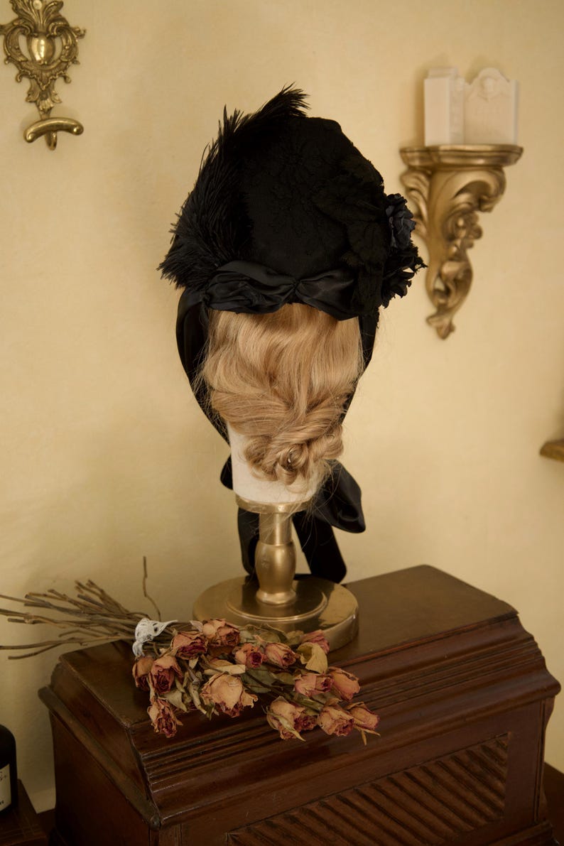 May include: A black vintage hat with feather and ribbon accents, displayed on a mannequin head with blonde hair. The hat is part of a vintage fashion collection. The display is on a gold stand, with dried flowers and a wooden cabinet in the background.