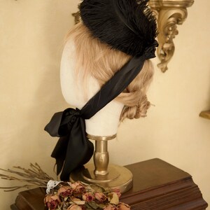 May include: A black feather fascinator with a satin ribbon, displayed on a mannequin head. The head is on a gold stand, with a bouquet of dried roses and branches on a wooden surface.