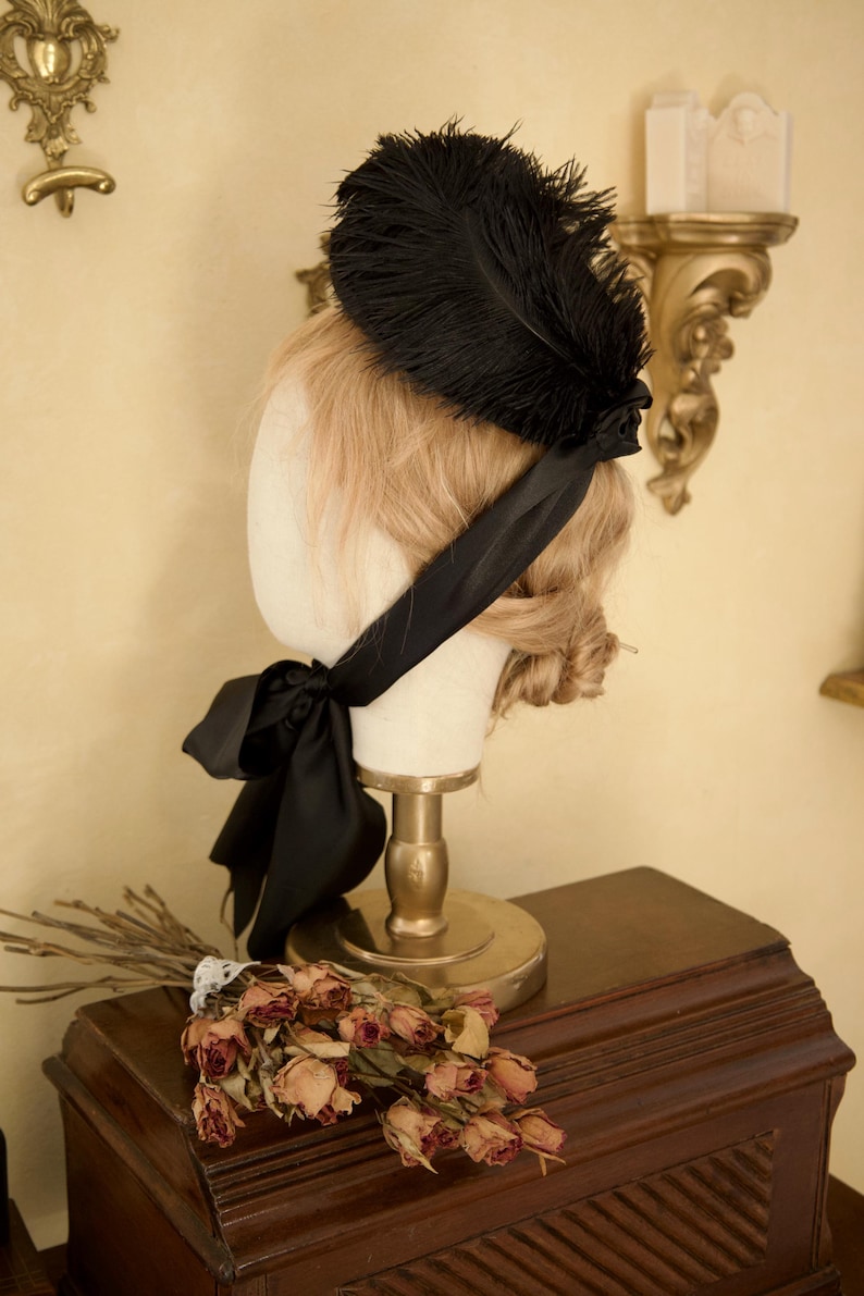 May include: A black feather fascinator with a satin ribbon, displayed on a mannequin head. The head is on a gold stand, with a bouquet of dried roses and branches on a wooden surface.