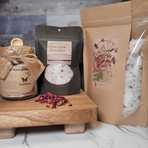 May include: Three bags of bath salts with labels that say "Divine Garden Botanical Bath". The bags are made of brown paper and have a clear plastic window to see the product inside. The bath salts are white with pink and purple accents. There are dried rose petals on a wooden surface in front of the bags.