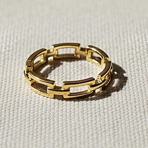 May include: A gold ring with a chain link design. The ring is made of a shiny, reflective metal and sits on a textured, off-white surface. The ring's design features rectangular links connected to each other.