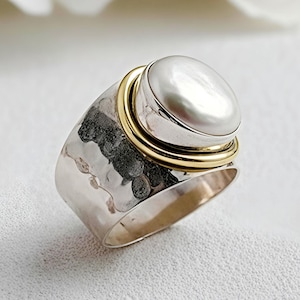 Könnte beinhalten: Ein Silberring mit einer großen, ovalen, weißen Perle, die in einem goldenen Band gefasst ist. Der Ring hat eine gehämmerten Textur auf dem Silberband. Die Perle ist der Blickfang und sorgt für einen klassischen und eleganten Look.