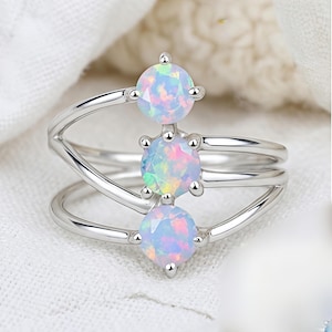 Può includere: Un anello d'argento con tre pietre preziose di opale iridescenti. L'anello ha un design unico con una fascia a strati e gli opali sono incastonati con griffe. Gli opali mostrano una gamma di colori, tra cui blu, rosa e verde.