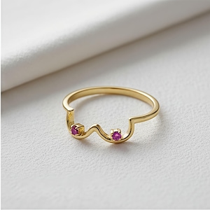 Könnte beinhalten: Ein goldfarbener Ring mit einem einzigartigen Design, das zwei kleine, runde, rote Edelsteine aufweist. Der Ring hat eine wellenförmige, asymmetrische Form, vor weißem Hintergrund.