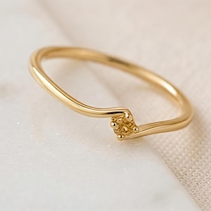 May include: A thin gold ring featuring a smooth, curved band that tapers toward a small, square-shaped setting. The setting contains a cluster of four tiny, textured gold prongs arranged in a square pattern at the center of the ring's curve.