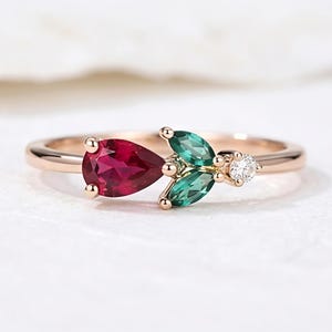 Könnte beinhalten: Ein Ring aus Roségold mit einem birnenförmigen roten Edelstein, zwei marquise-geschliffenen grünen Edelsteinen in Blattform und einem kleinen runden weißen Diamanten, gefasst auf einer polierten Roségoldschiene mit glatter Oberfläche.