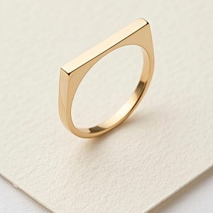 May include: A gold ring featuring a rectangular bar design. The ring, made of shiny metal, is set against a white backdrop. The band is smooth and rounded, complementing the bar's straight lines. A piece of jewellery.