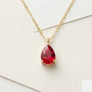May include: A gold chain necklace featuring a pear-shaped, faceted deep red gemstone pendant. The pendant is secured in a gold prong setting, suspended from a delicate gold link chain that connects to the top of the teardrop-shaped stone.