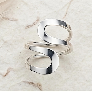 May include: A silver-colored ring with a unique, open design. The ring features a swirling, abstract shape with smooth, polished surfaces. The ring is displayed on a textured, off-white surface, showcasing its modern aesthetic.