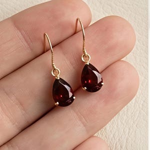 May include: Gold-coloured drop earrings with teardrop-shaped, deep red gemstones. The earrings have a simple hook design. The faceted gemstones catch the light, adding sparkle. Jewellery.