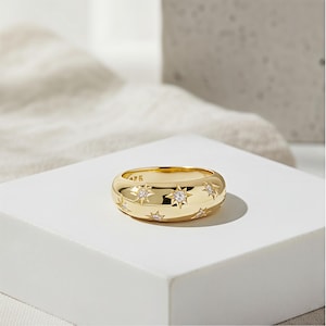 May include: A gold-colored ring with a rounded band. The ring is adorned with star-shaped designs, each featuring a central, sparkling stone. The ring is displayed on a white surface, with a soft, neutral background.