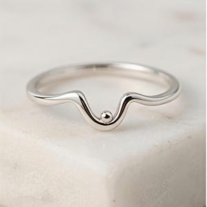 Puede incluir: Un anillo de tono plateado pulido que presenta una banda lisa con forma de onda. El centro del anillo muestra una pequeña cuenta esférica situada en el punto más bajo de la curva, creando un diseño limpio y sencillo con acabado brillante.